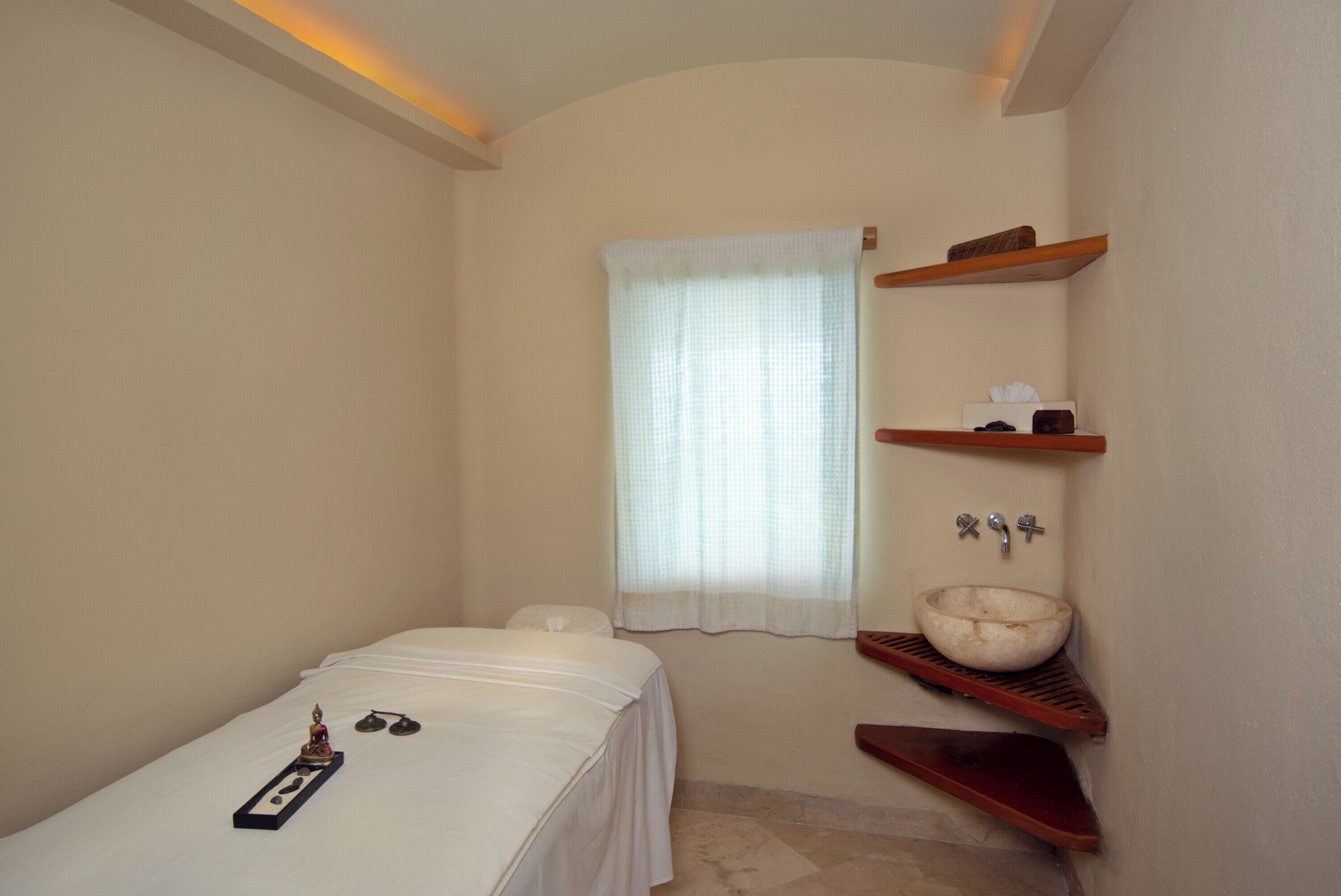 Une petite chambre avec un lit simple, une table de chevet, une fenêtre avec des rideaux légers, une étagère en bois où sont rangés les articles de toilette et un lavabo intégré.