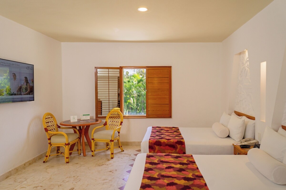 Une chambre d’hôtel avec un lit, une petite table et des chaises, une fenêtre avec des stores et un tableau au mur.