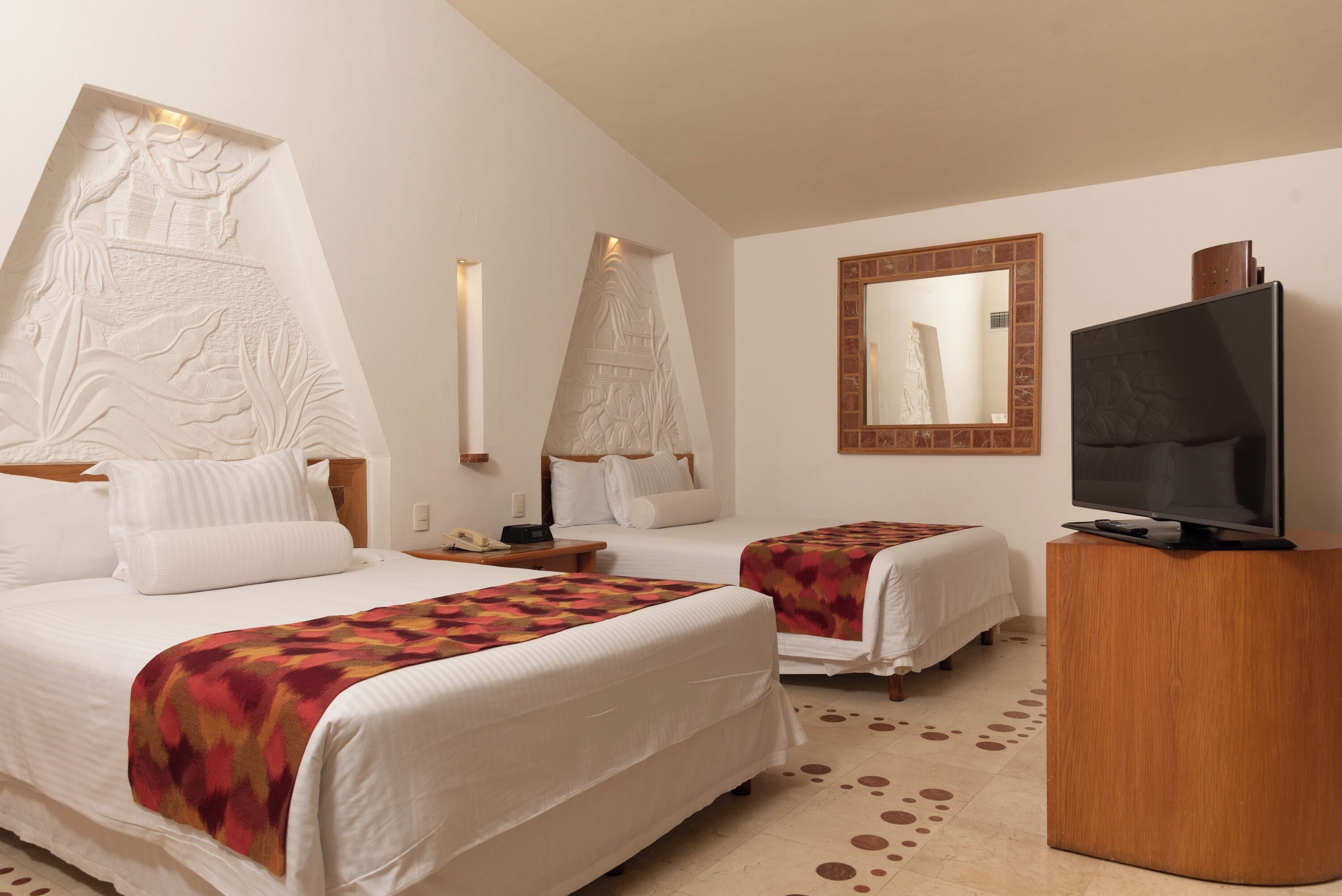 A hotel room with two beds, a TV, a mirror, and decorative wall carvings.
