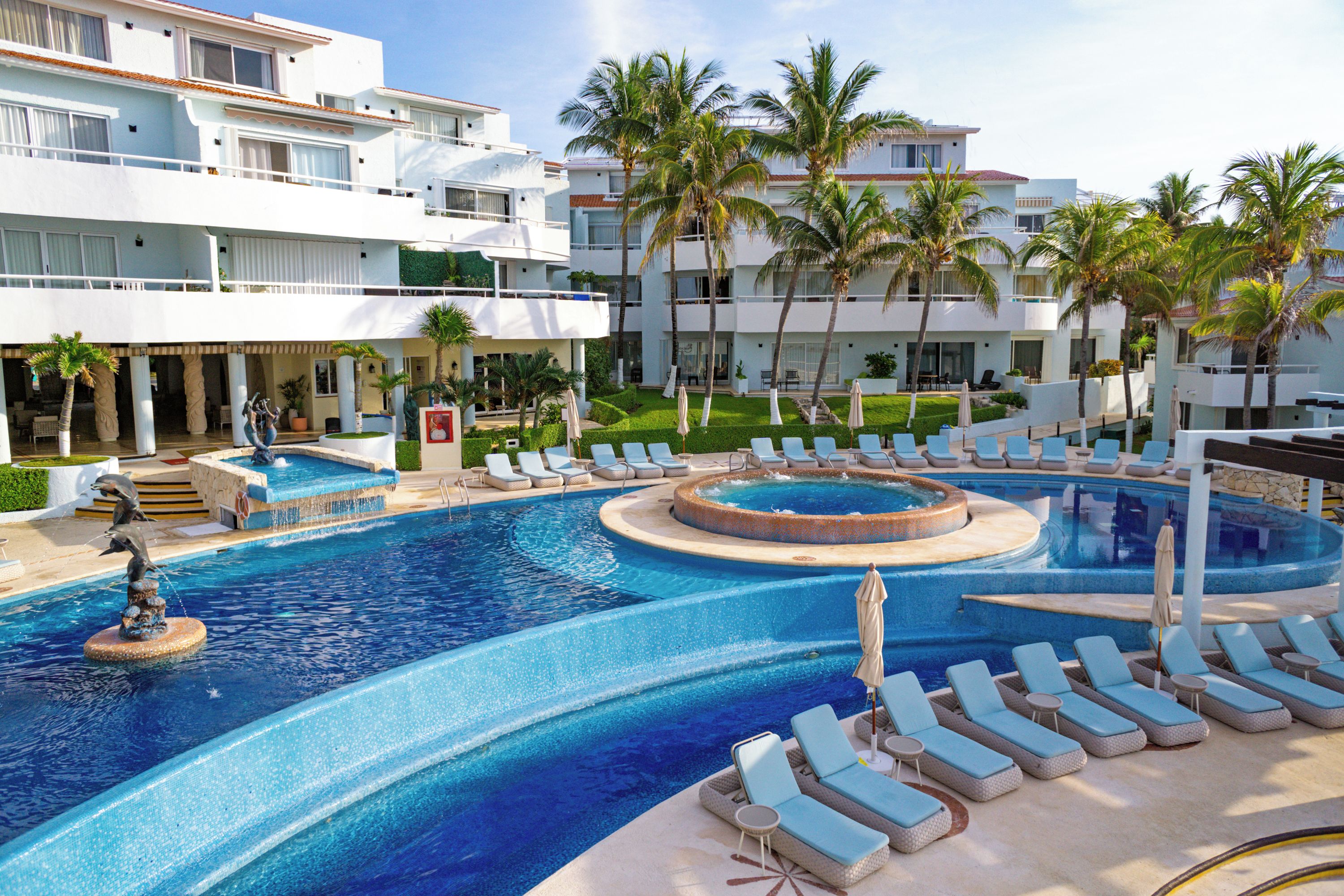 Een resort met een groot zwembad, ligbedden, palmbomen en een gebouw met balkons.