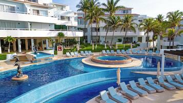 Een resort met een groot zwembad, ligbedden, palmbomen en een gebouw met balkons.