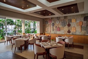 A restaurant with multiple tables and chairs, a stone wall with various kitchen utensils, and large windows overlooking a tropical garden. - Sunset Fishermen Beach Resort Playa del Carmen - All inclusive (Playa del Carmen)