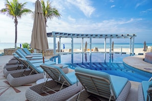 A poolside area with lounge chairs and umbrellas, a pergola, and a view of the beach.
