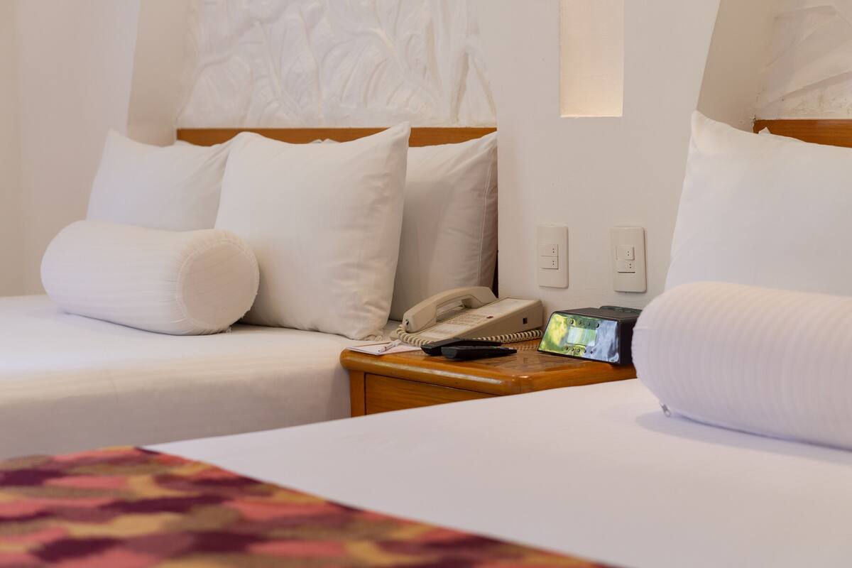 Ein Hotelzimmer mit zwei Betten, einem Nachttisch mit Telefon und Fernbedienung sowie einer gemusterten Bettdecke.