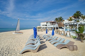 A beach with lounge chairs, a sailboat, and a building with a pool.