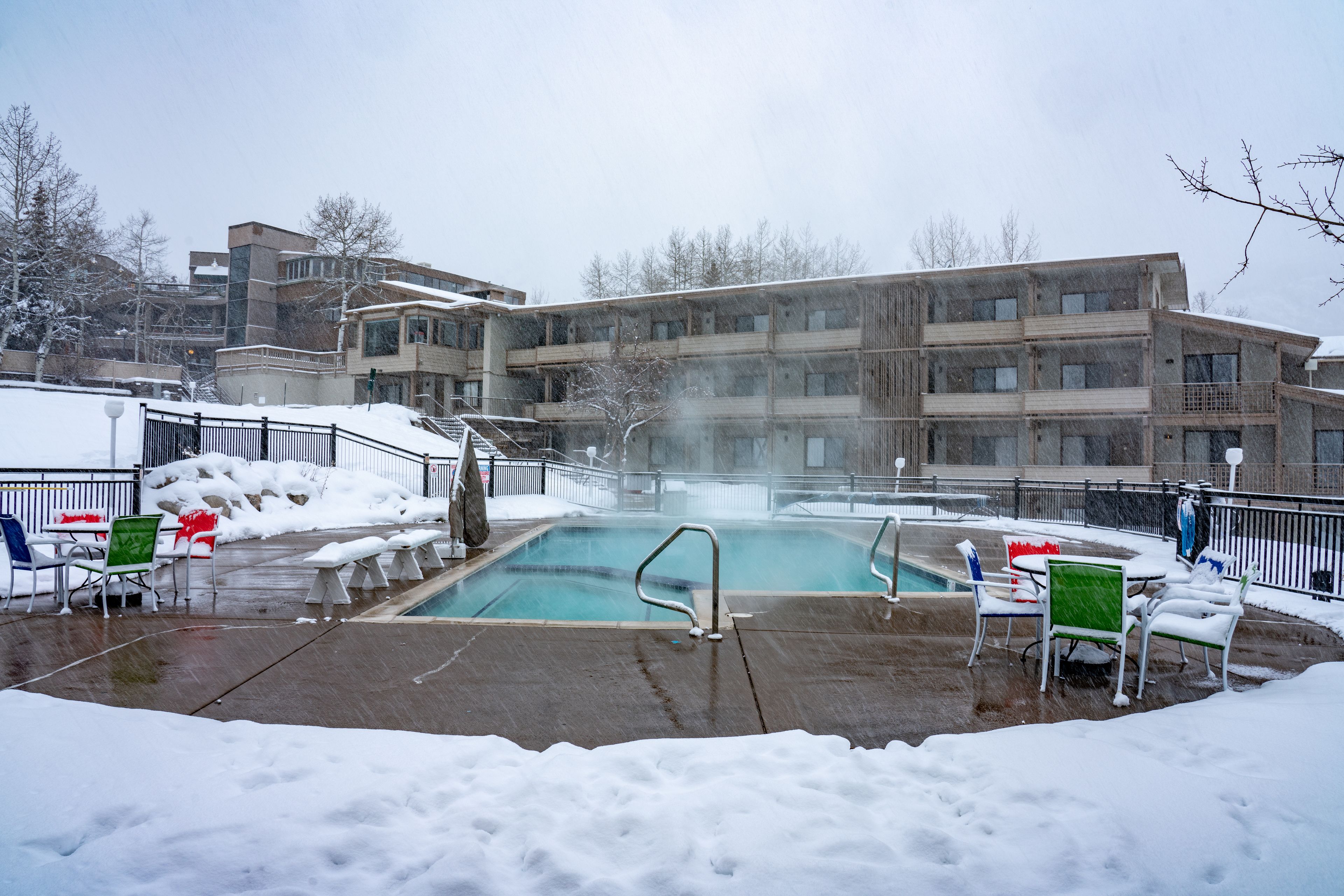 A pool area with a hot tub, surrounded by snow-covered ground and a multi-story building in the background.