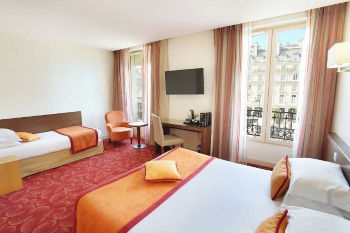 Habitación de hotel con dos camas, un escritorio, una silla, un televisor y una ventana con vistas a edificios.