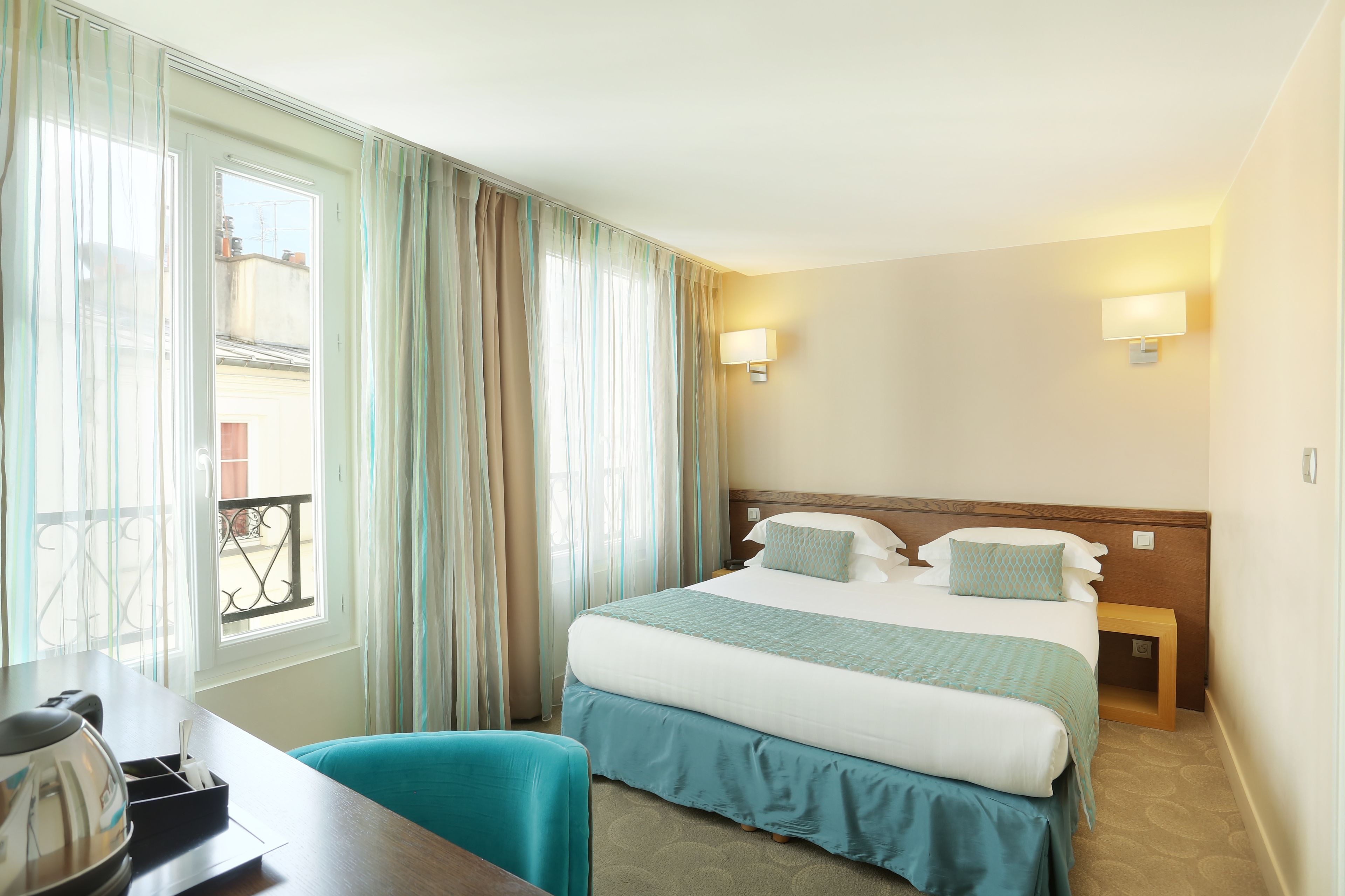 Habitación de hotel con cama, mesitas de noche, escritorio con hervidor y vistas al exterior.