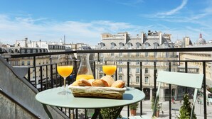 Ein Balkon mit Frühstückstisch, einschließlich Brot und Orangensaft, mit Blick auf eine Stadtlandschaft mit historischen Gebäuden.