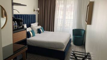 Ein Hotelzimmer mit Bett, Schreibtisch, Stuhl, Mülleimer und einem Fenster mit Vorhängen.