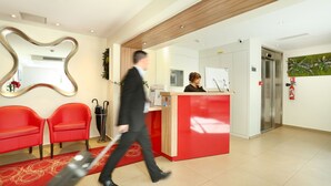 Ein Hotel-Rezeptionsbereich mit einem roten Empfangstresen, zwei roten Stühlen, einem großen Spiegel und einem Feuerlöscher an der Wand.