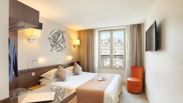 Ein Hotelzimmer mit Bett, Schreibtisch mit Laptop, Stuhl, Fernseher und Fenster mit Blick auf Gebäude.
