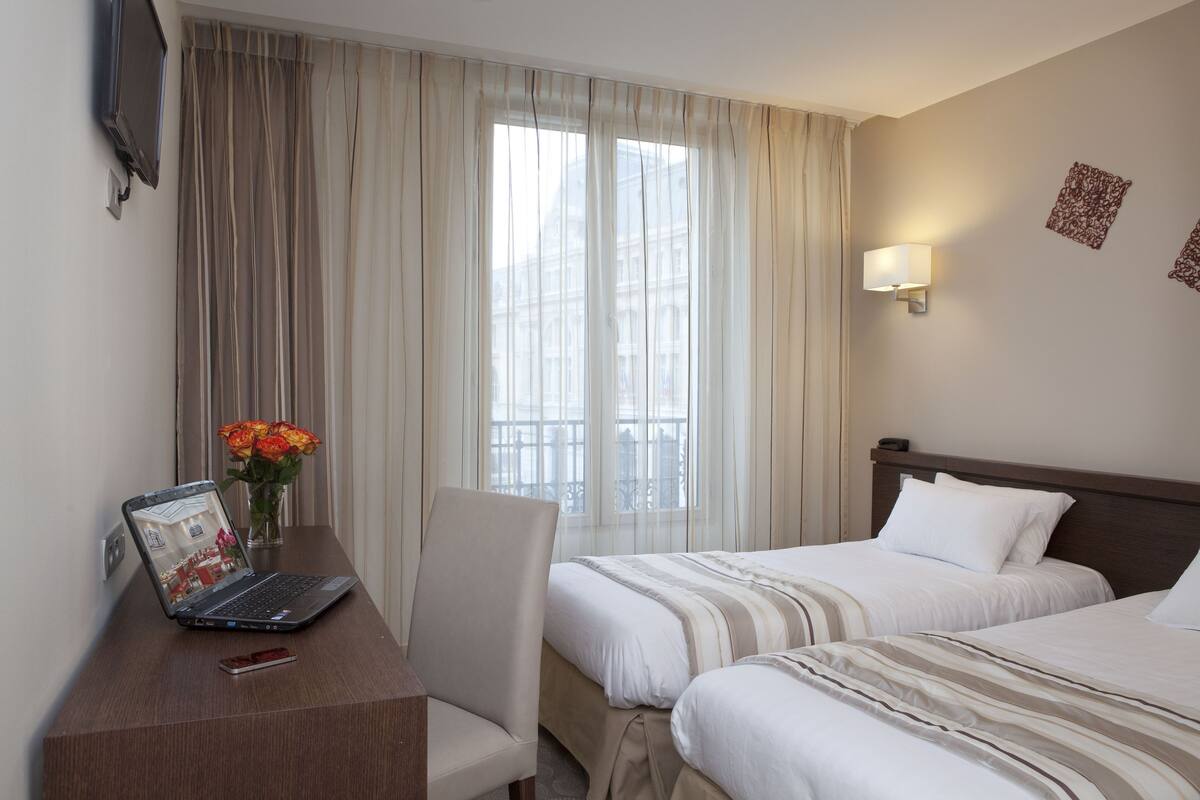 Hotellihuone, jossa on kaksi sänkyä, työpöytä kannettavalla tietokoneella, tuoli, maljakko kukkineen ja ikkuna, jossa on verhot.