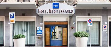 A hotel entrance with a sign reading "HOTEL MEDITERRANEO". There are flags above the entrance, a parking sign, and a no parking sign.