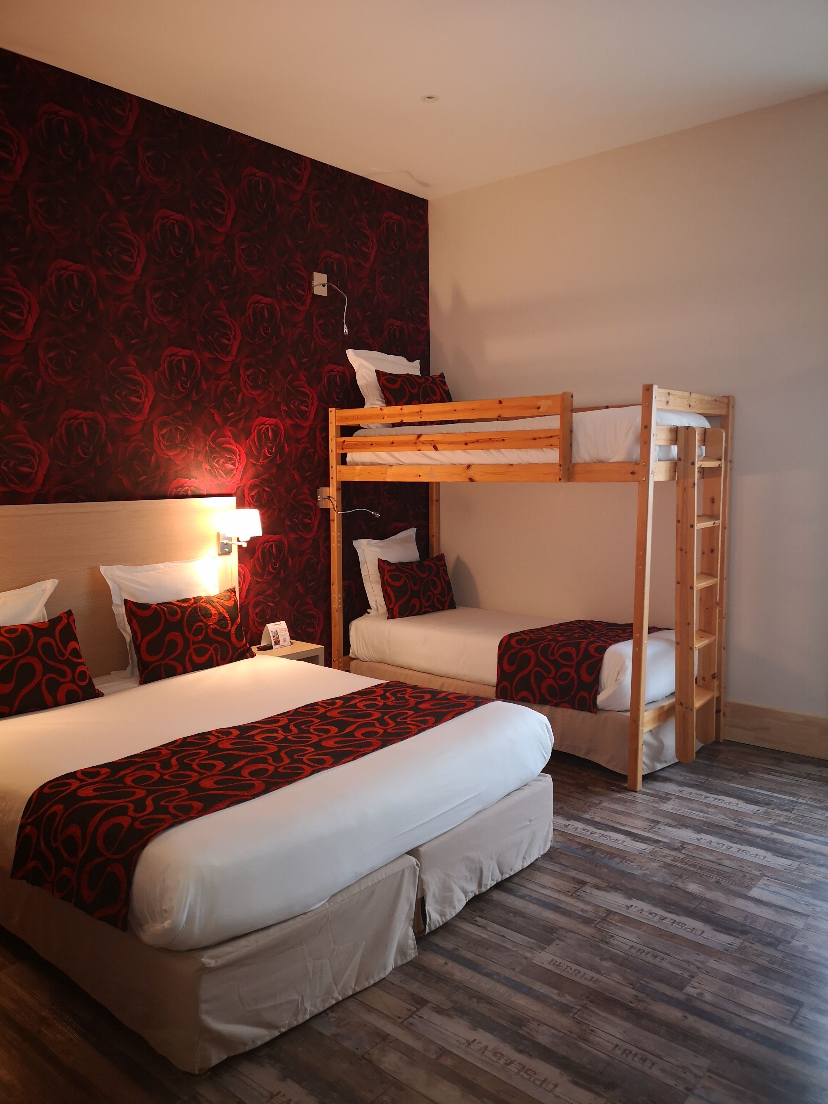 Une chambre avec un lit superposé, un papier peint à motifs de roses rouges, du linge de lit blanc et un plancher en bois.