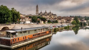 A riverboat docked near a historic building with a tower, surrounded by a calm river and urban buildings.