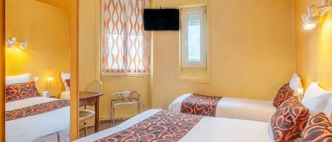 A hotel room with two beds, a desk, a chair, a TV mounted on the wall, and a window with patterned curtains.