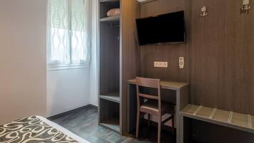 Una habitación con cama, un armario de madera, un escritorio con silla, un televisor de pantalla plana montado en la pared y una ventana con cortinas estampadas.