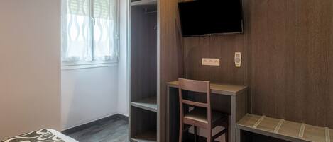 A room with a bed, a wooden wardrobe, a desk with a chair, a flat-screen TV mounted on the wall, and a window with patterned curtains.