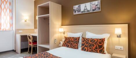 A hotel room with a bed, two bedside lamps, a desk with a chair, a wardrobe, and a wall-mounted picture of the Eiffel Tower.