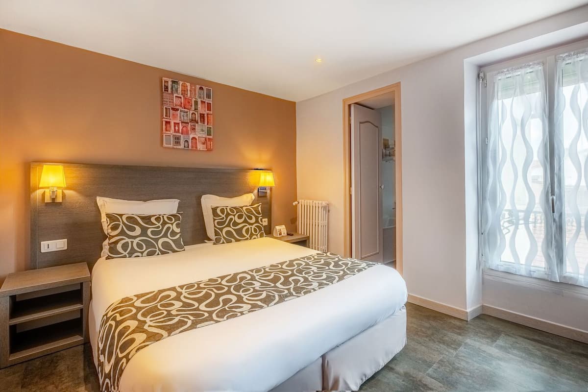 Een hotelkamer met een groot bed, twee bedlampen, een radiator, een deur naar de badkamer en een raam met gordijn.