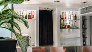 Un coin bar avec des étagères remplies de bouteilles d’alcool et de verres, un rideau sombre et une plante au premier plan.