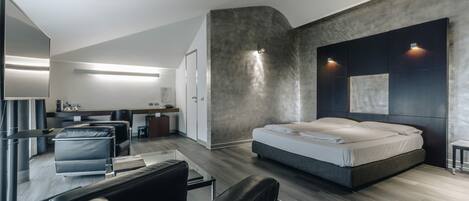 A modern hotel room with a large bed, a seating area with a glass table, and a kitchenette in the background.