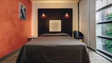 Quarto de hotel moderno com uma cama grande, uma mesinha de cabeceira pequena, uma cadeira, uma janela ampla com vista para o verde e um aplique de parede.