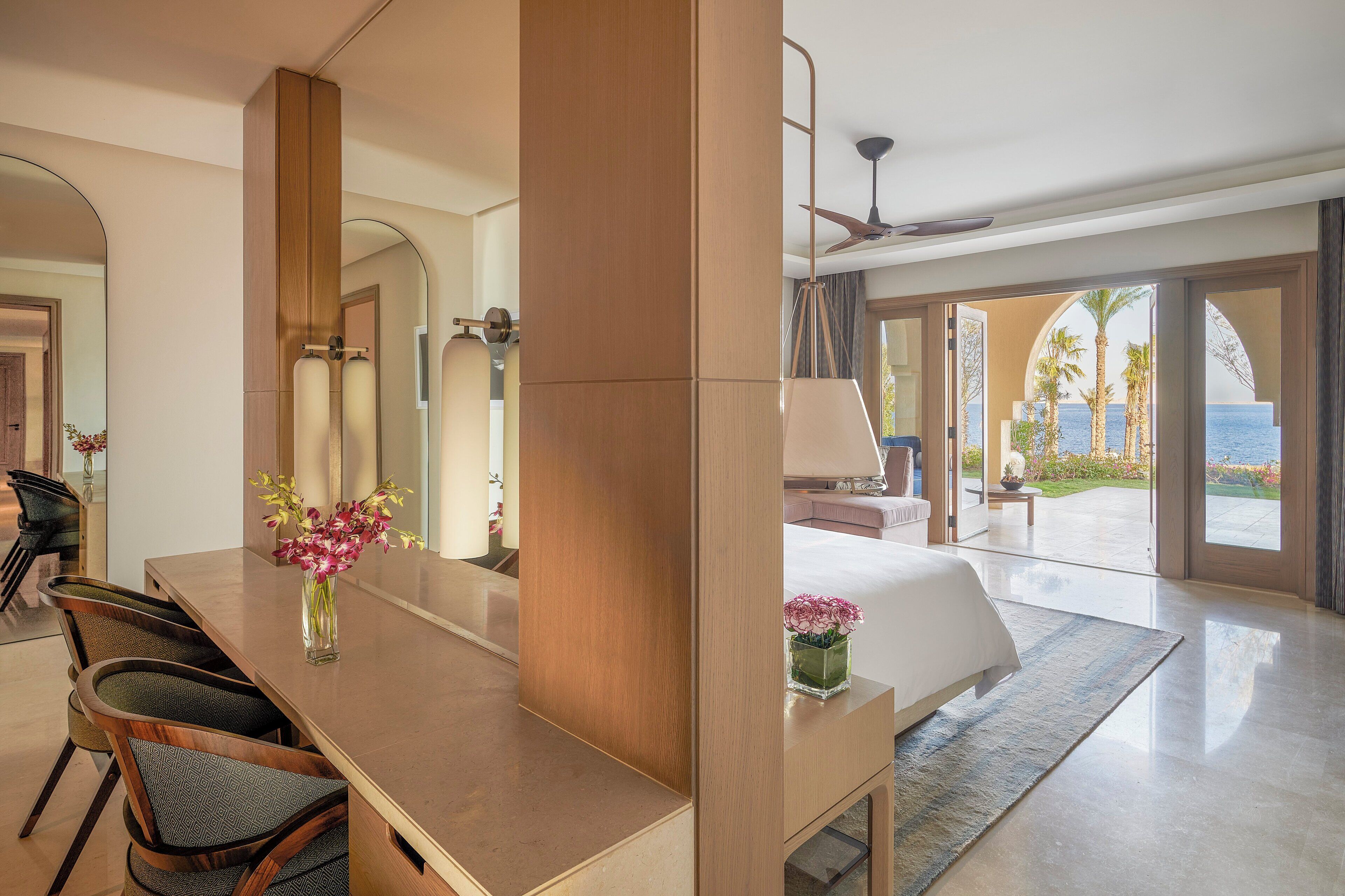 A modern hotel room with a large bed, a dining area with chairs, a small table with flowers, and a view of the ocean through large glass doors.