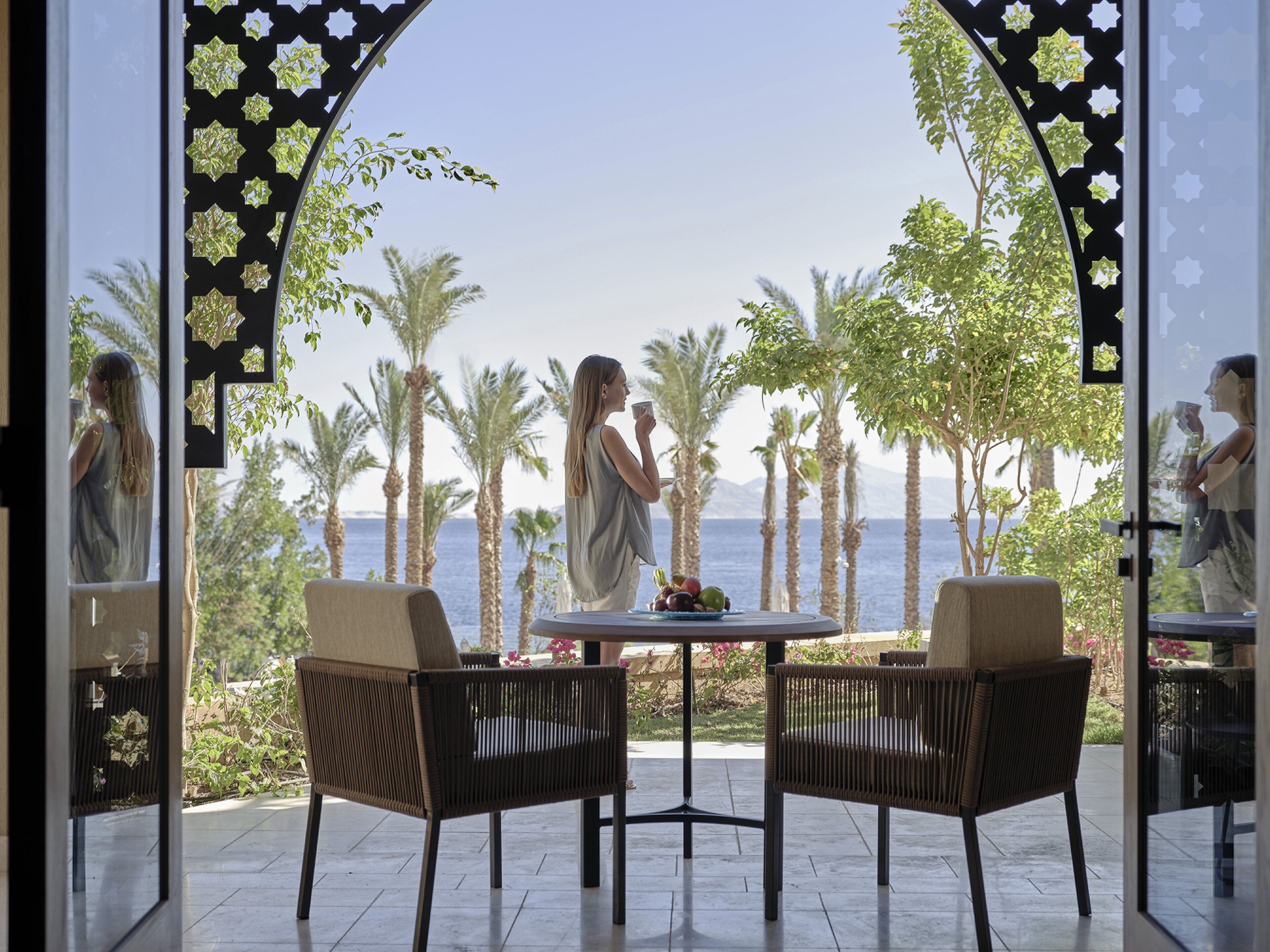 A room with a view of the sea, two wicker chairs, and a table with fruit.