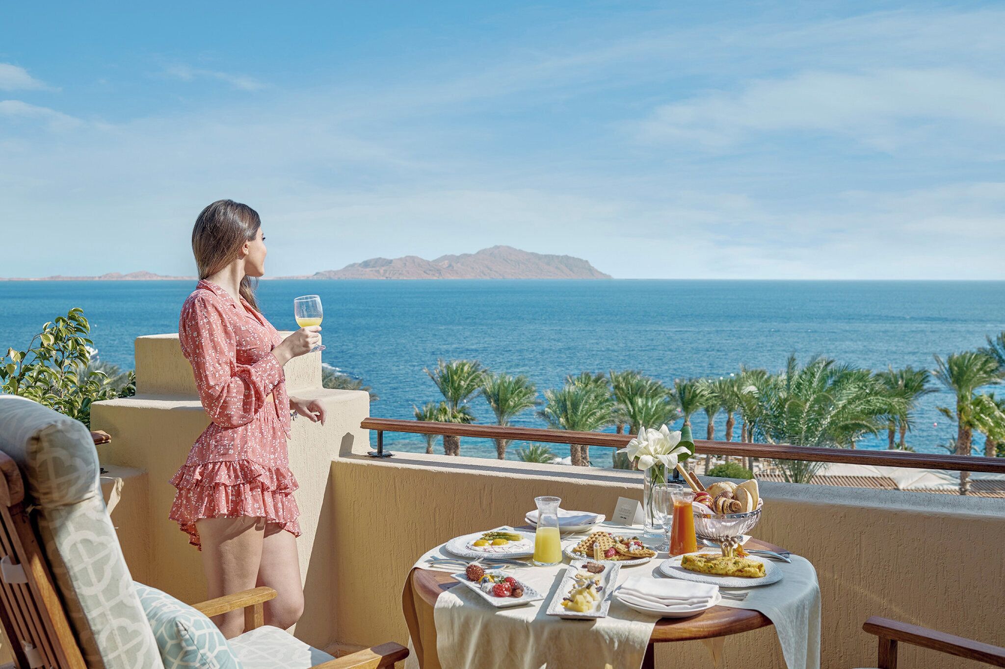 Eine Frau genießt eine Mahlzeit auf einem Balkon mit Blick auf das Meer und Palmen.