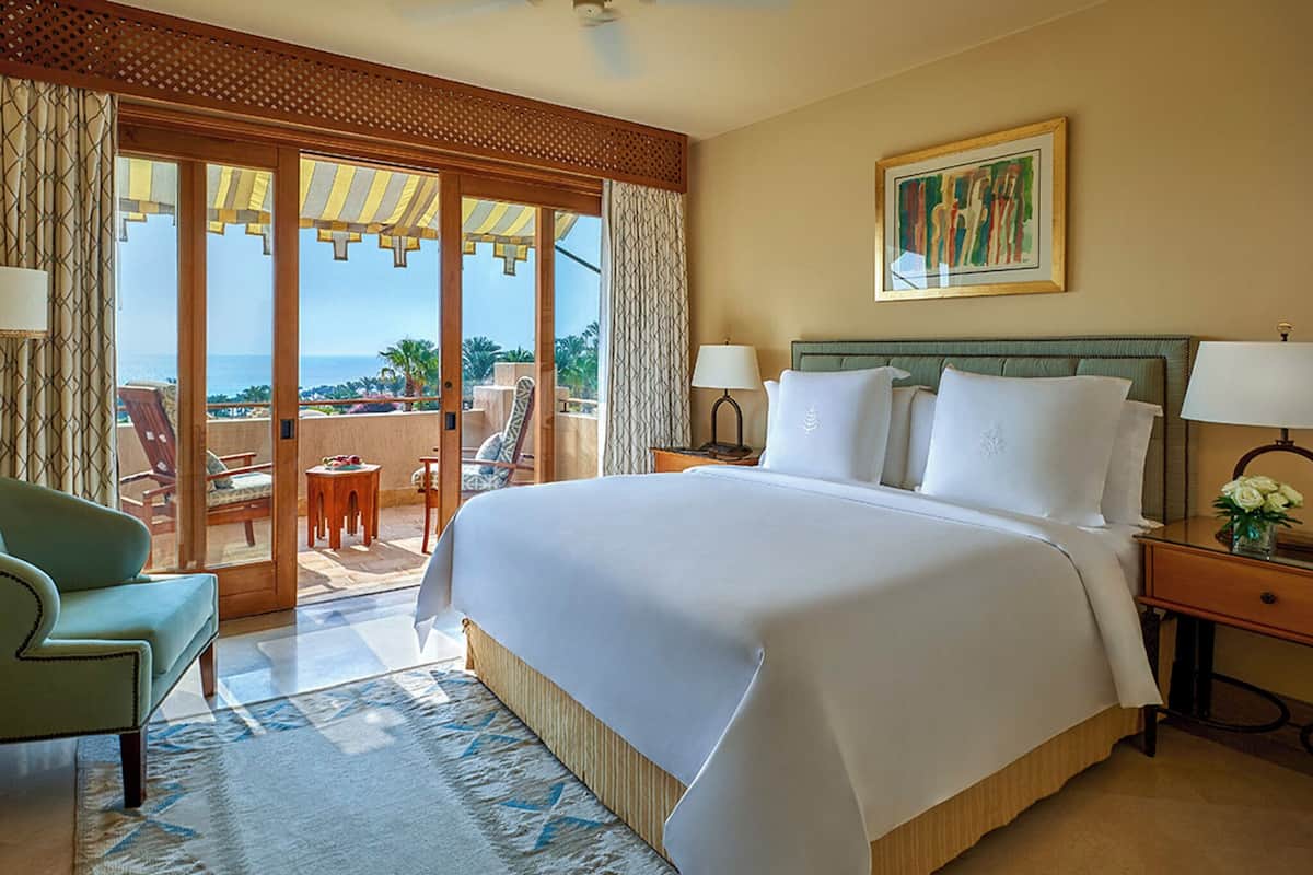 A hotel room with a large bed, a chair, a small table, and a view of the ocean through a sliding door.
