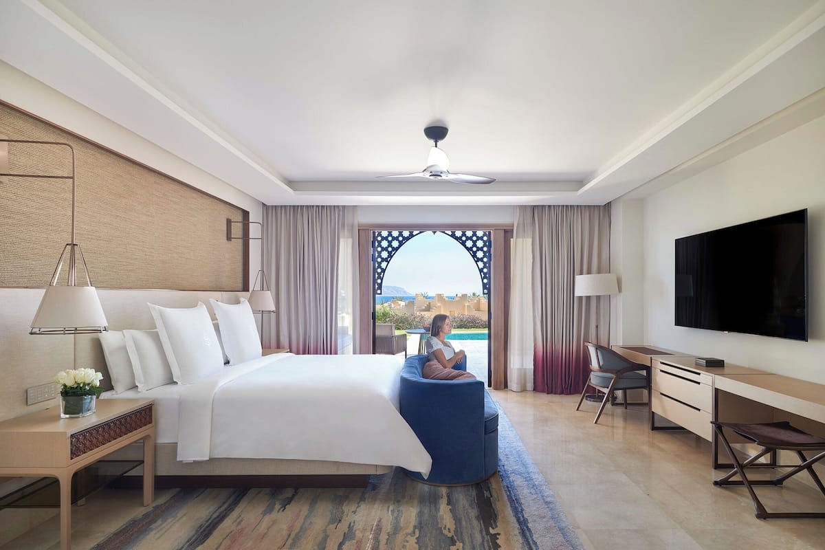 A modern hotel room with a large bed, a desk, a TV, and a view of a pool and mountains.
