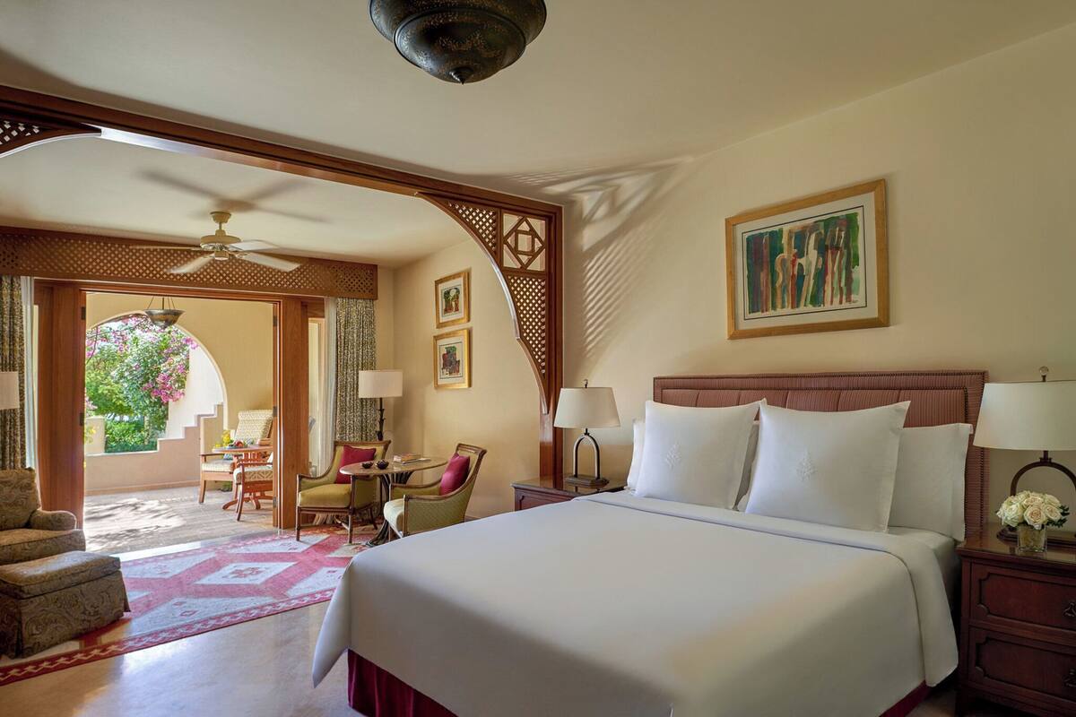 A hotel room with a large bed, a desk with a chair, a small table, and a view of a garden through an open doorway.