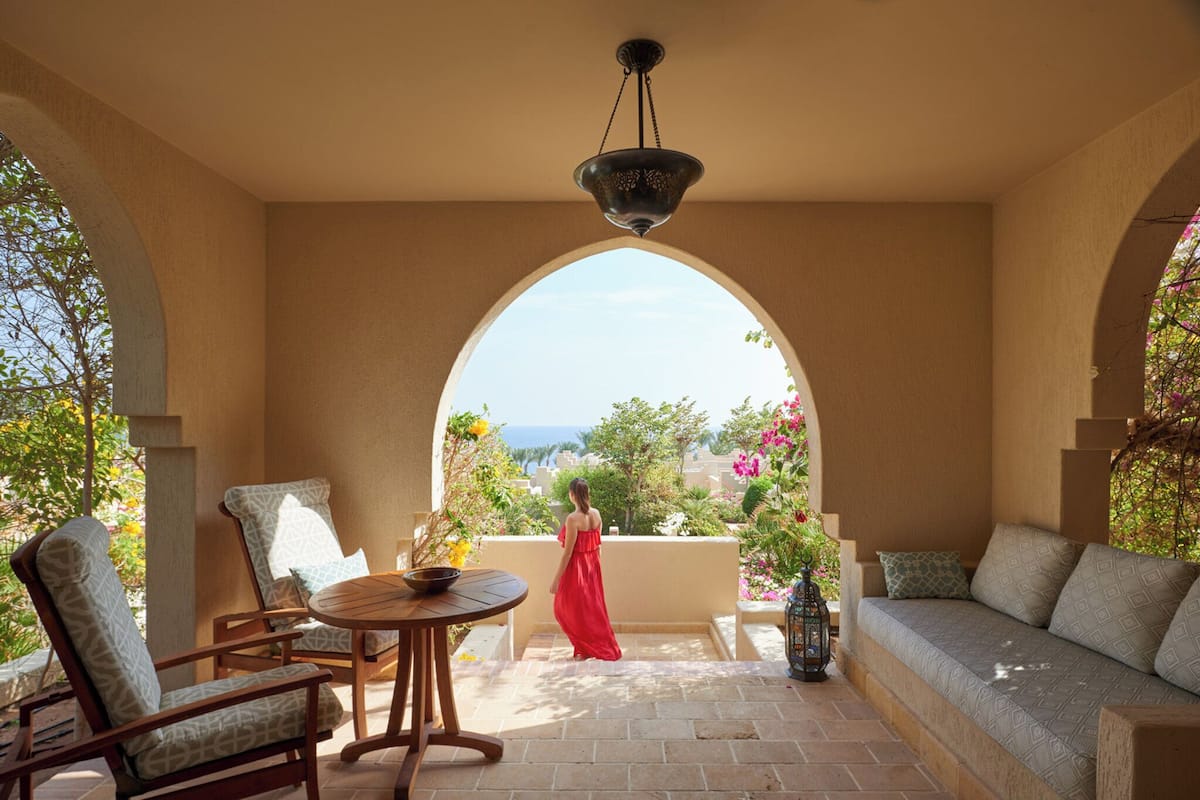 A sunlit patio with a view of the sea, featuring a seating area with a sofa and chairs, a table, and a hanging light fixture.
