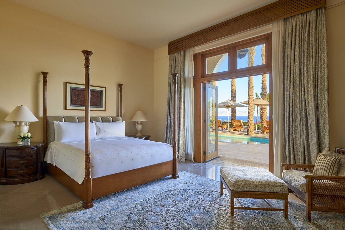 A spacious bedroom with a large bed, a sitting area with a chair, and a view of a pool and outdoor seating area.