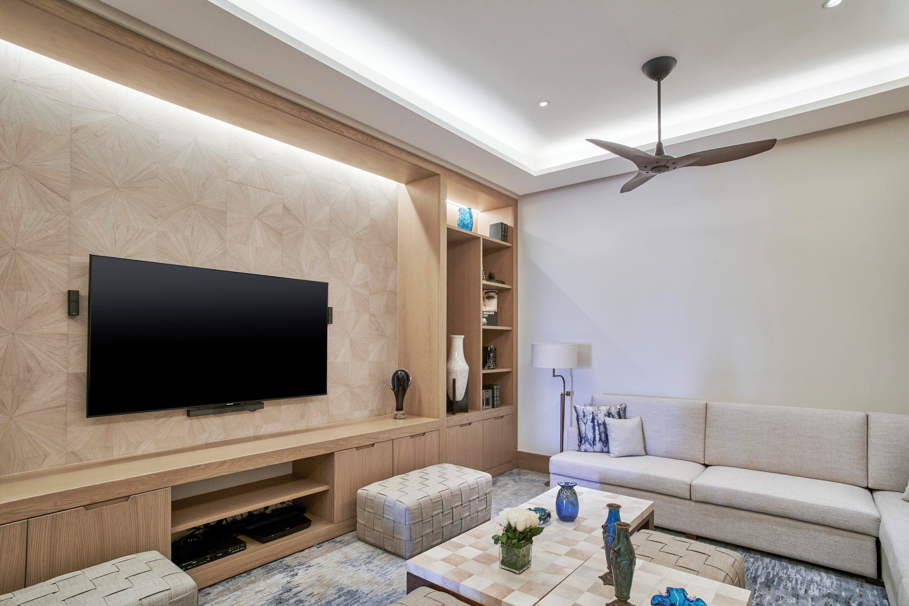 Un salon moderne doté d’un téléviseur à écran plat, d’étagères en bois, d’un ventilateur de plafond, d’un canapé et d’une table basse agrémentée d’objets décoratifs.