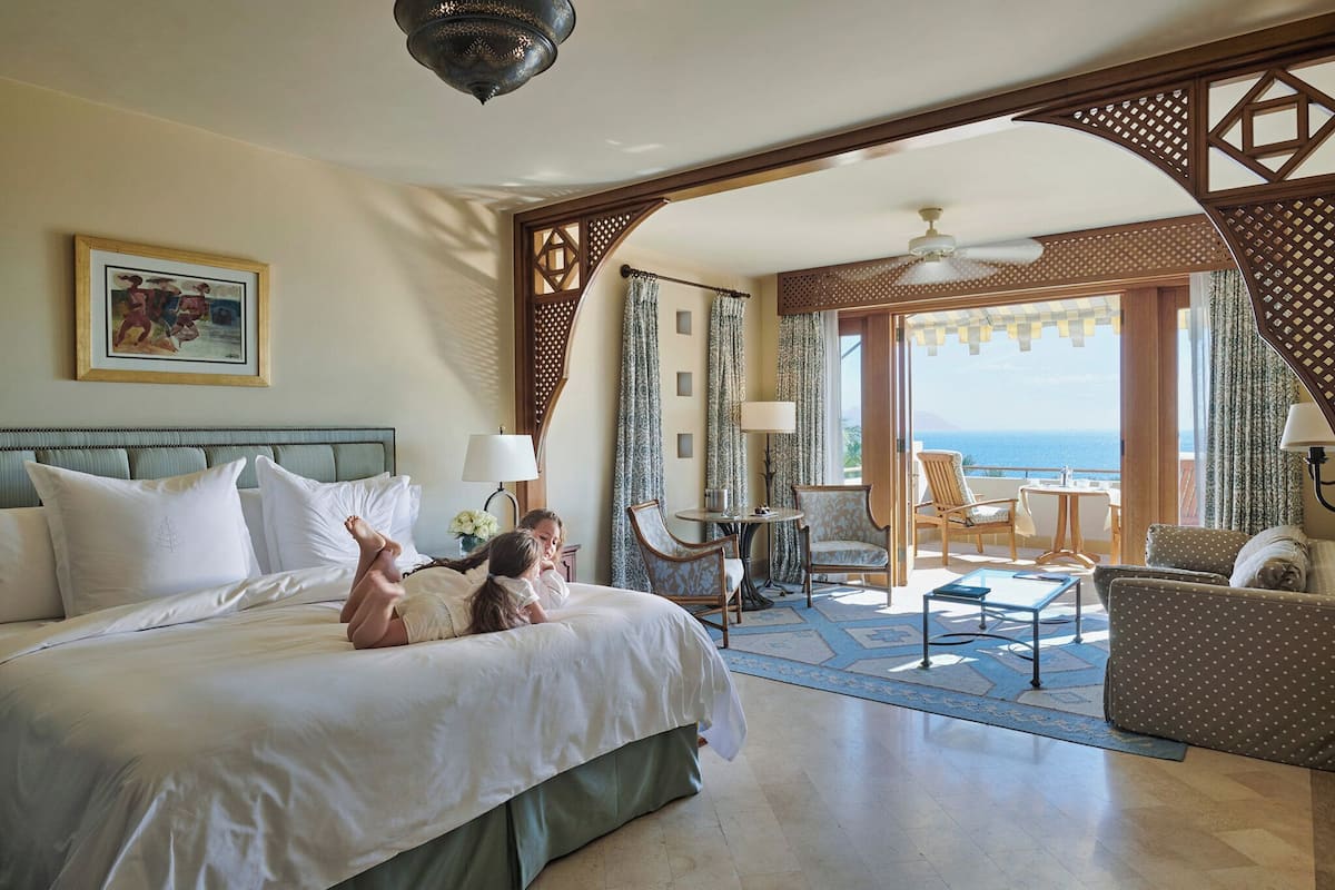A hotel room with a large bed, two chairs, a small table, and a view of the sea through a balcony.