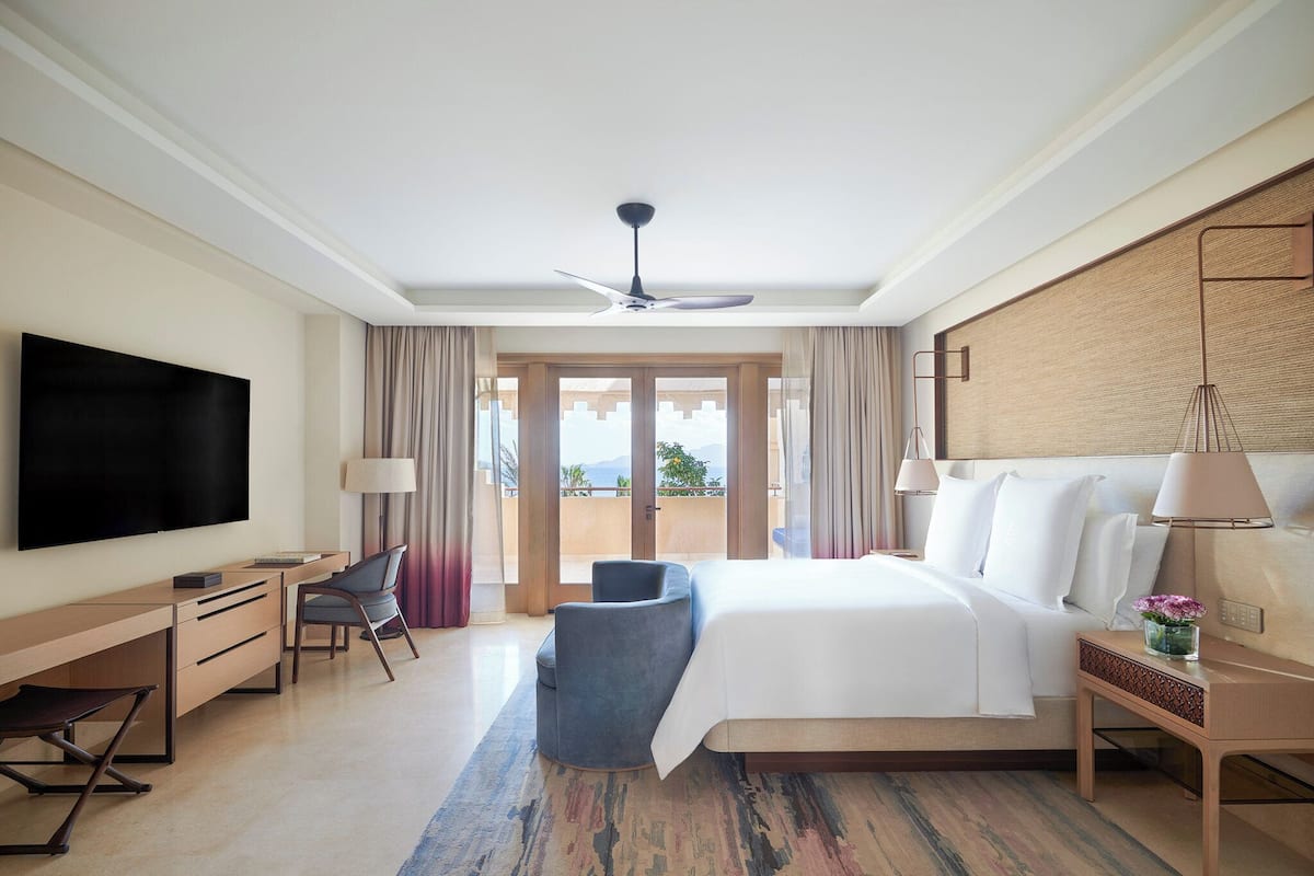 A hotel room with a large bed, a desk, a chair, a TV, and a balcony with curtains.