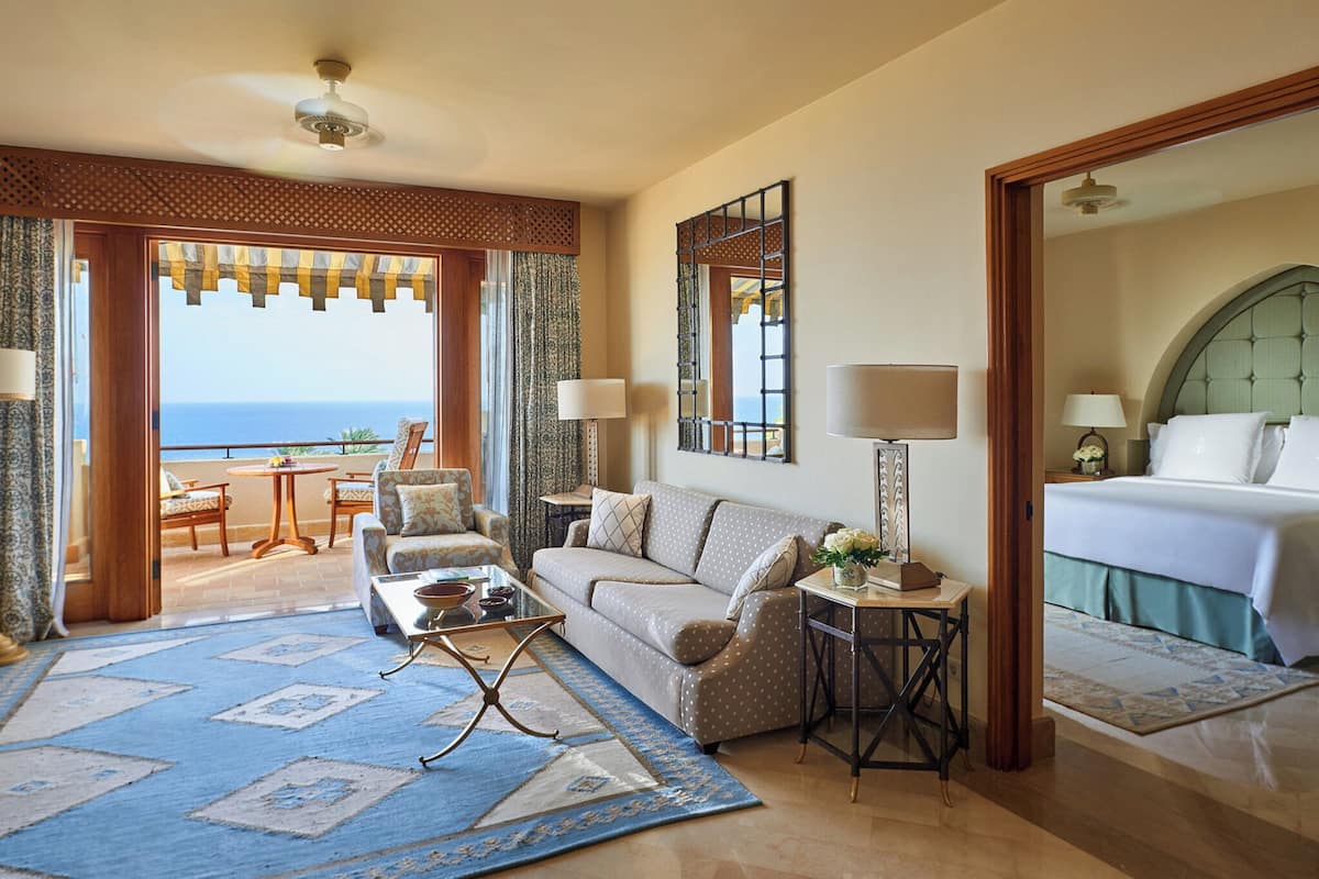 A hotel room with a view of the sea, a sofa, a coffee table, a side table with a lamp, and a bed with a headboard.