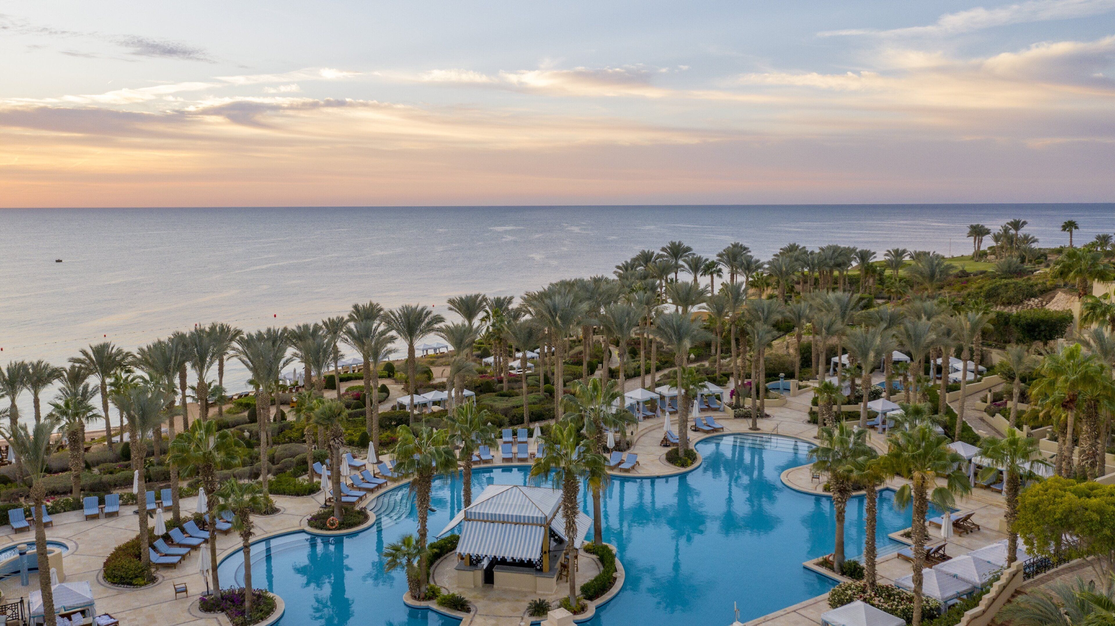 Een resort met een groot zwembad, palmbomen en uitzicht op zee.