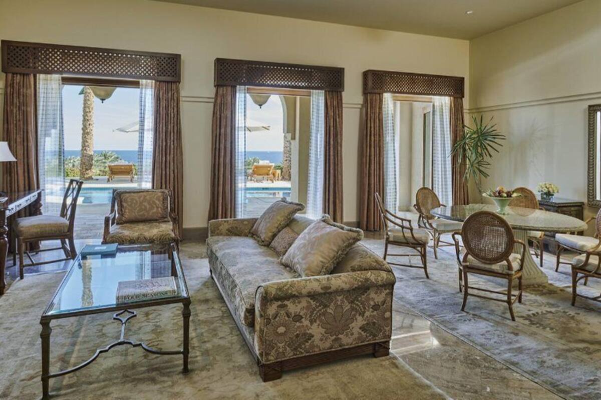 A spacious living room with a sofa, armchairs, a glass coffee table, and a dining area with wicker chairs. Large windows offer a view of a pool and outdoor seating.