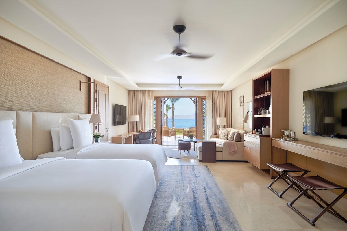 A hotel room with two beds, a desk, a sofa, a TV, and a view of the sea.