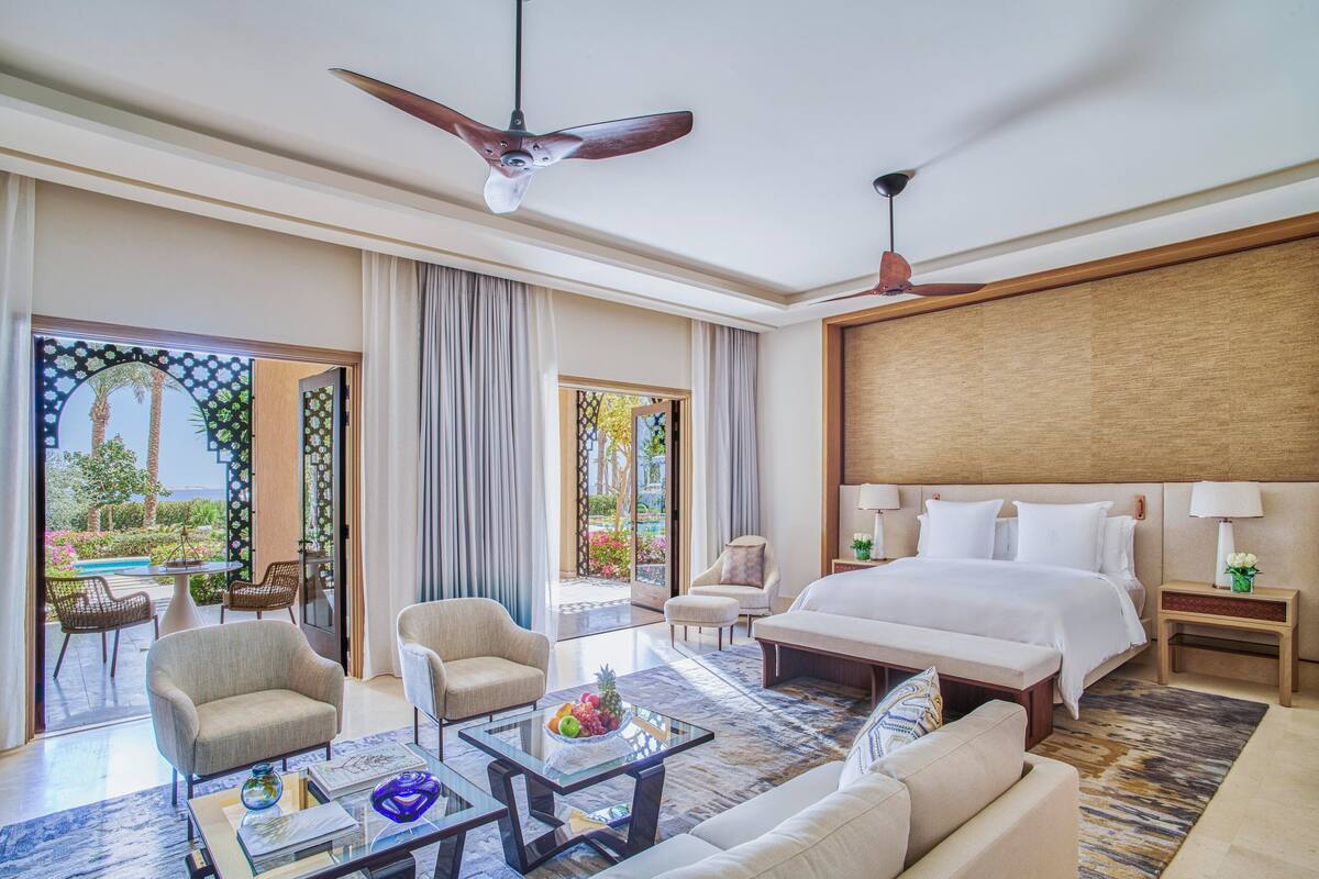 A spacious hotel room with a large bed, two armchairs, a coffee table, and a view of a garden through a sliding door.