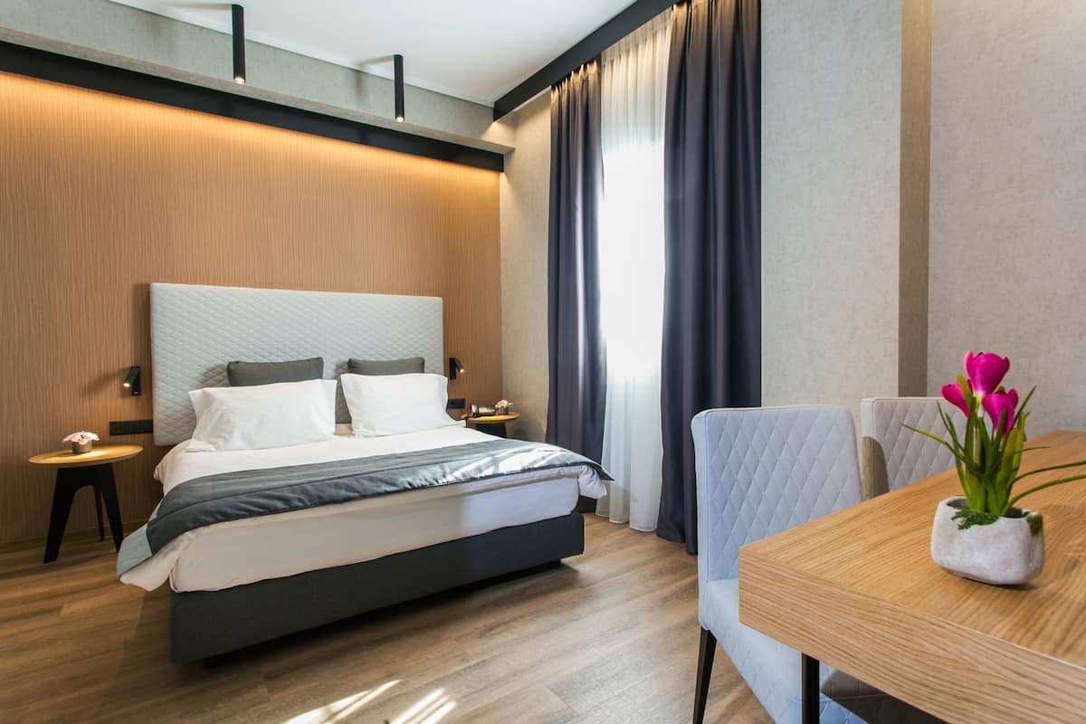 Ein modernes Hotelzimmer mit einem großen Bett, einem Holznachttisch, einem kleinen Tisch mit Blumen und Blick durch große Fenster nach draußen.