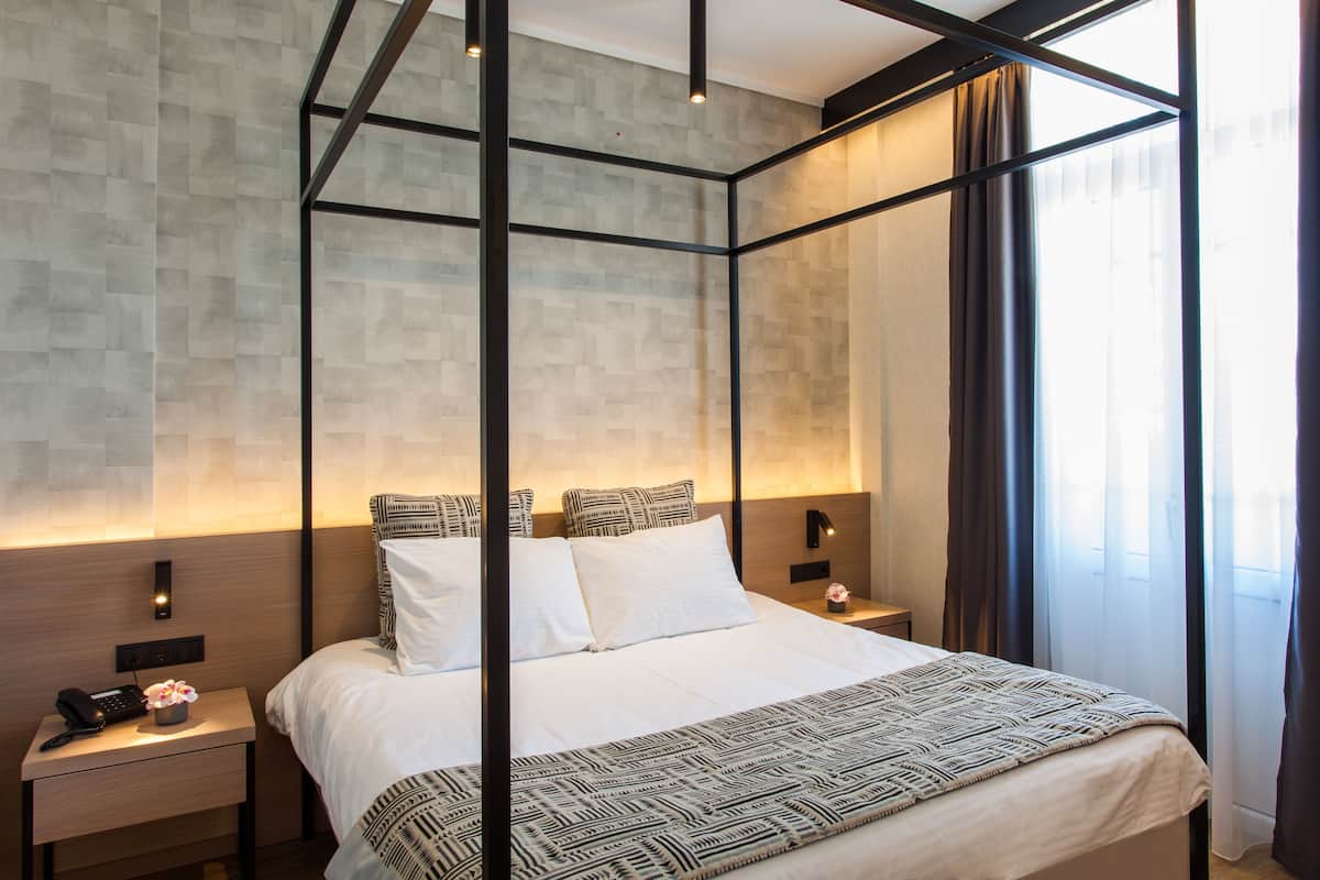 Ein modernes Hotelzimmer mit Himmelbett, Nachttisch mit Lampe und Blick auf die Stadt durch große Fenster.