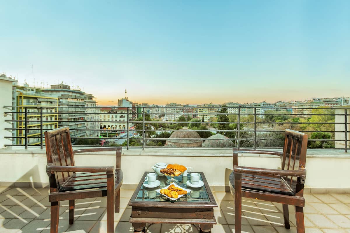 Eine Terrasse mit zwei Holzstühlen und einem Tisch, gedeckt für Tee, mit Blick auf eine Stadtlandschaft aus Gebäuden und Grünflächen.
