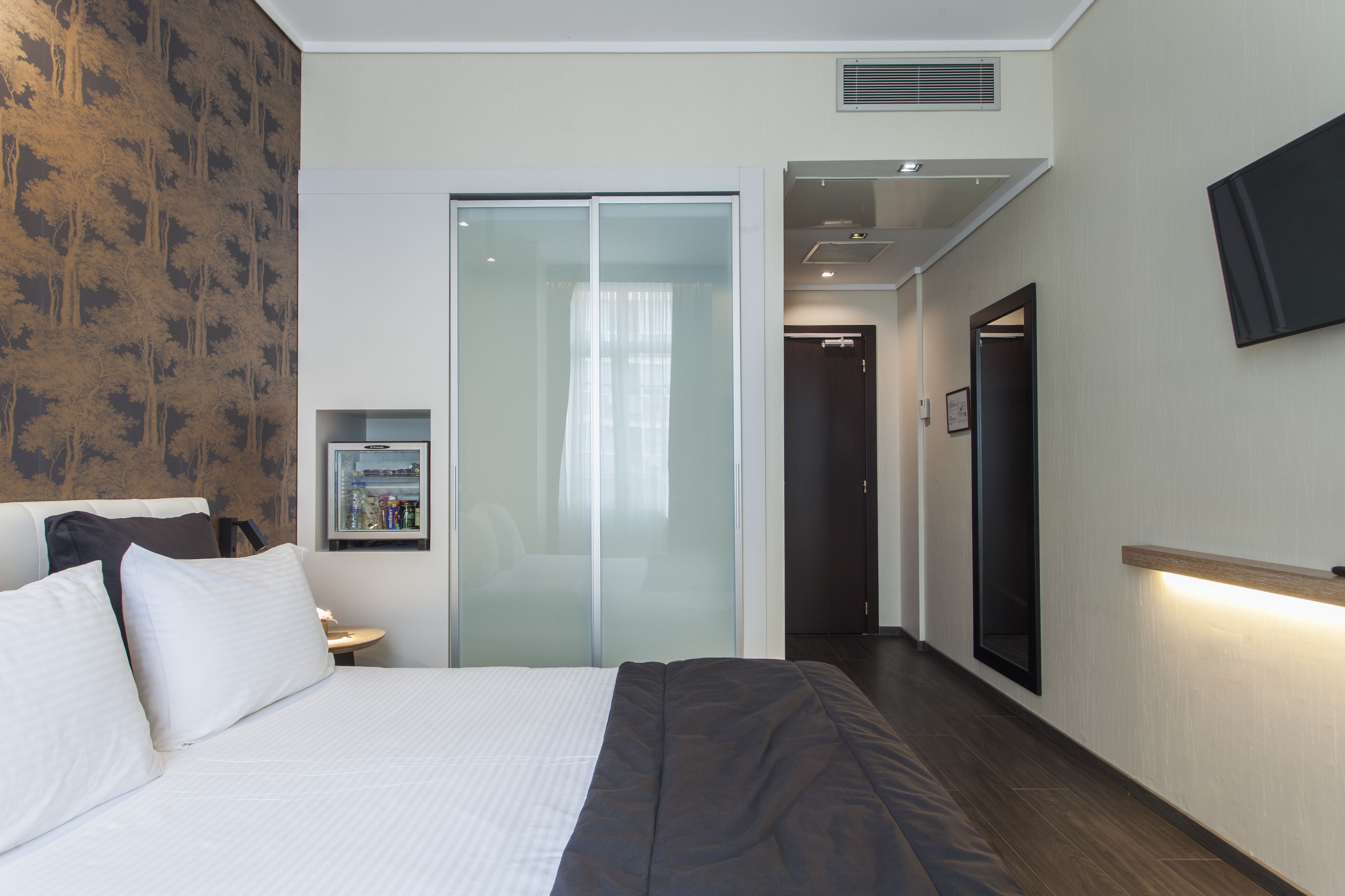 Ein modernes Hotelzimmer mit einem großen Bett, einem Schrank mit Spiegel, einem an der Wand befestigten Fernseher und einem kleinen Tisch mit Flasche und Gläsern.