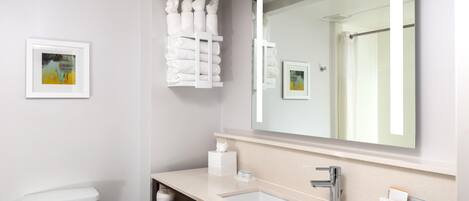 A modern bathroom with a toilet, sink, and large mirror. There are towels hanging on a rack and a framed picture on the wall.