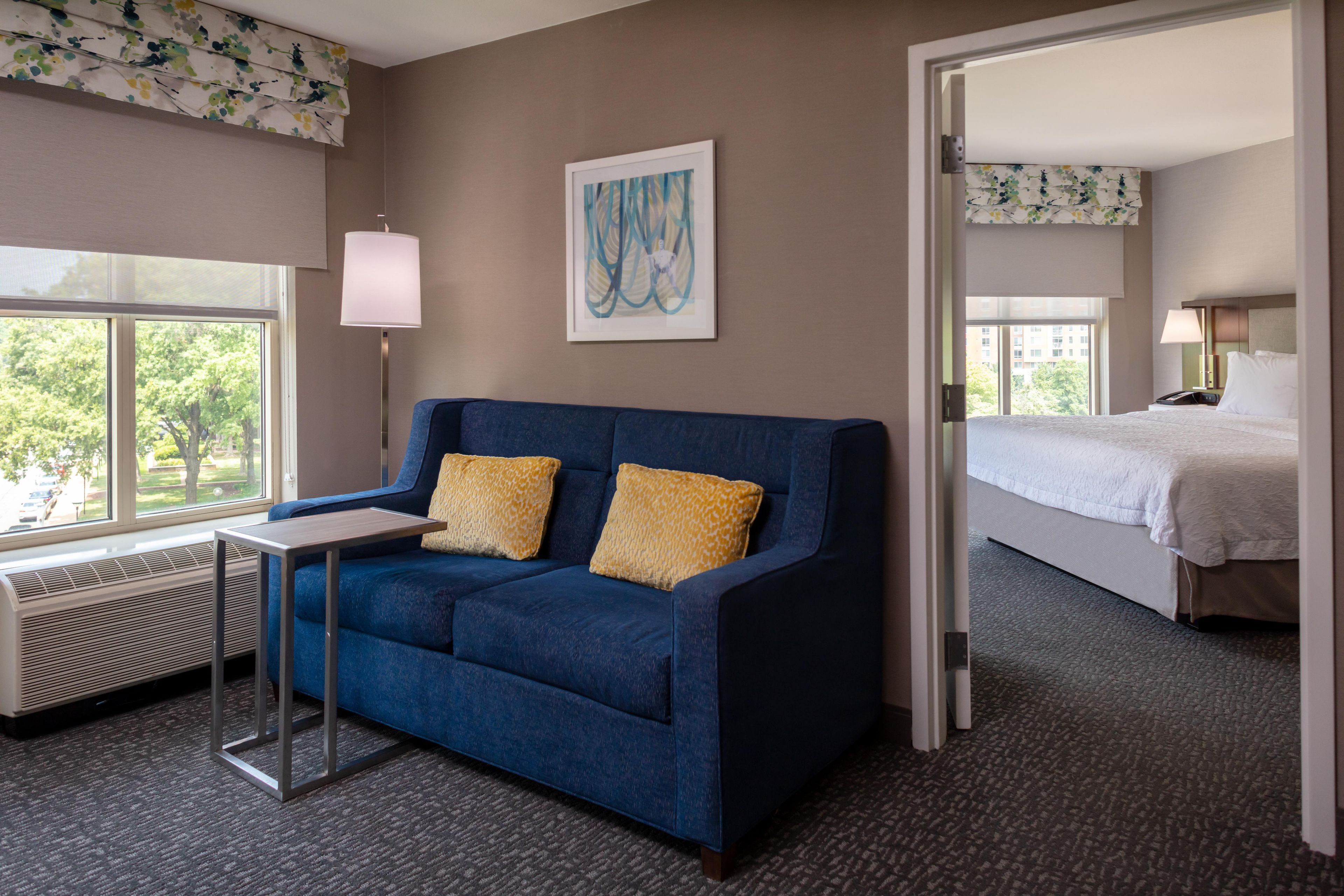 Een hotelkamer met een blauwe bank, een tafeltje, een lamp, een schilderij en een bed.